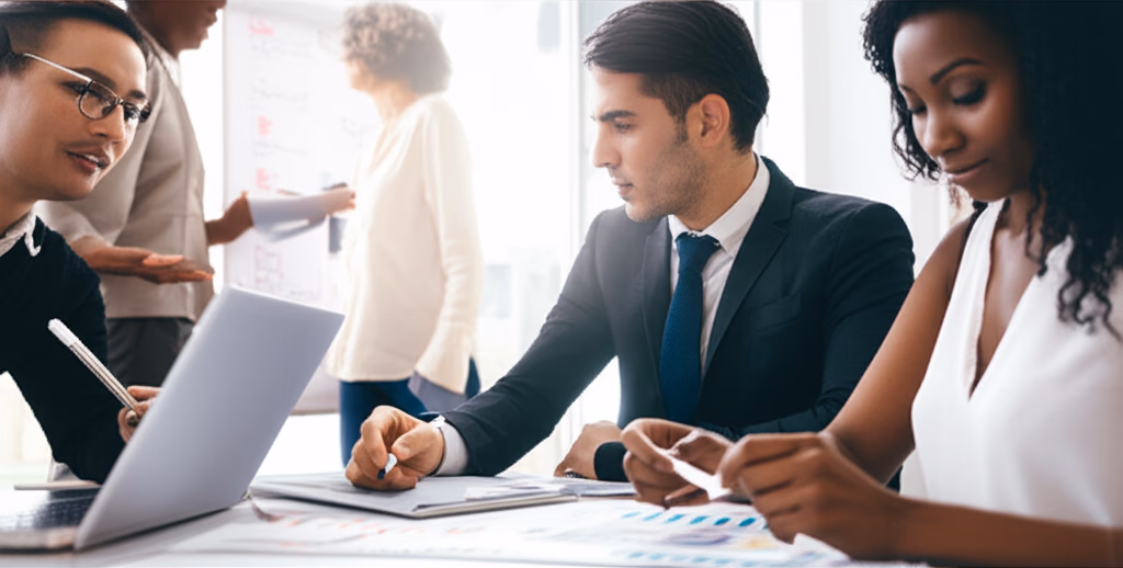 Diverse team of professionals actively collaborating around a table with laptops and a whiteboard, illustrating effective teamwork and brainstorming in a modern office.