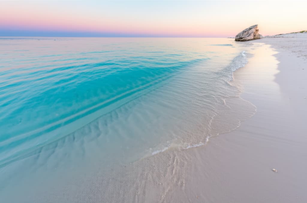 Ultra-realistic image of a tranquil tropical ocean view at sunrise with clear turquoise water, soft waves, and a pristine white sand beach under a pastel sky. Perfect for calming coastal scenes.
