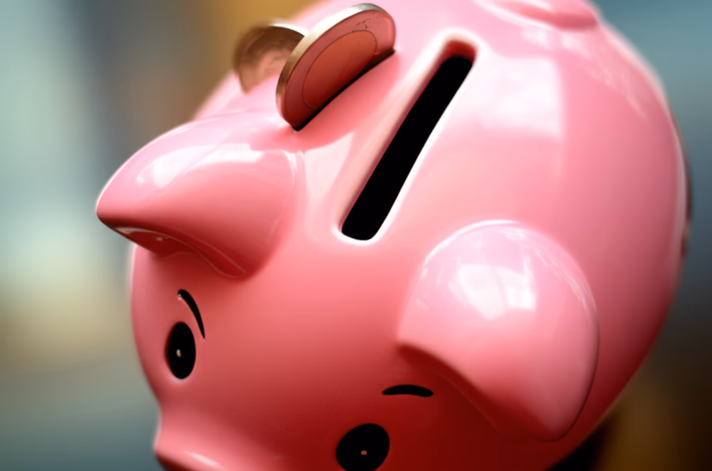 A traditional pink ceramic piggy bank with a coin slot on its back, sitting on a wooden surface, symbolizing saving money and financial security. A few shiny coins are visible in the slot.